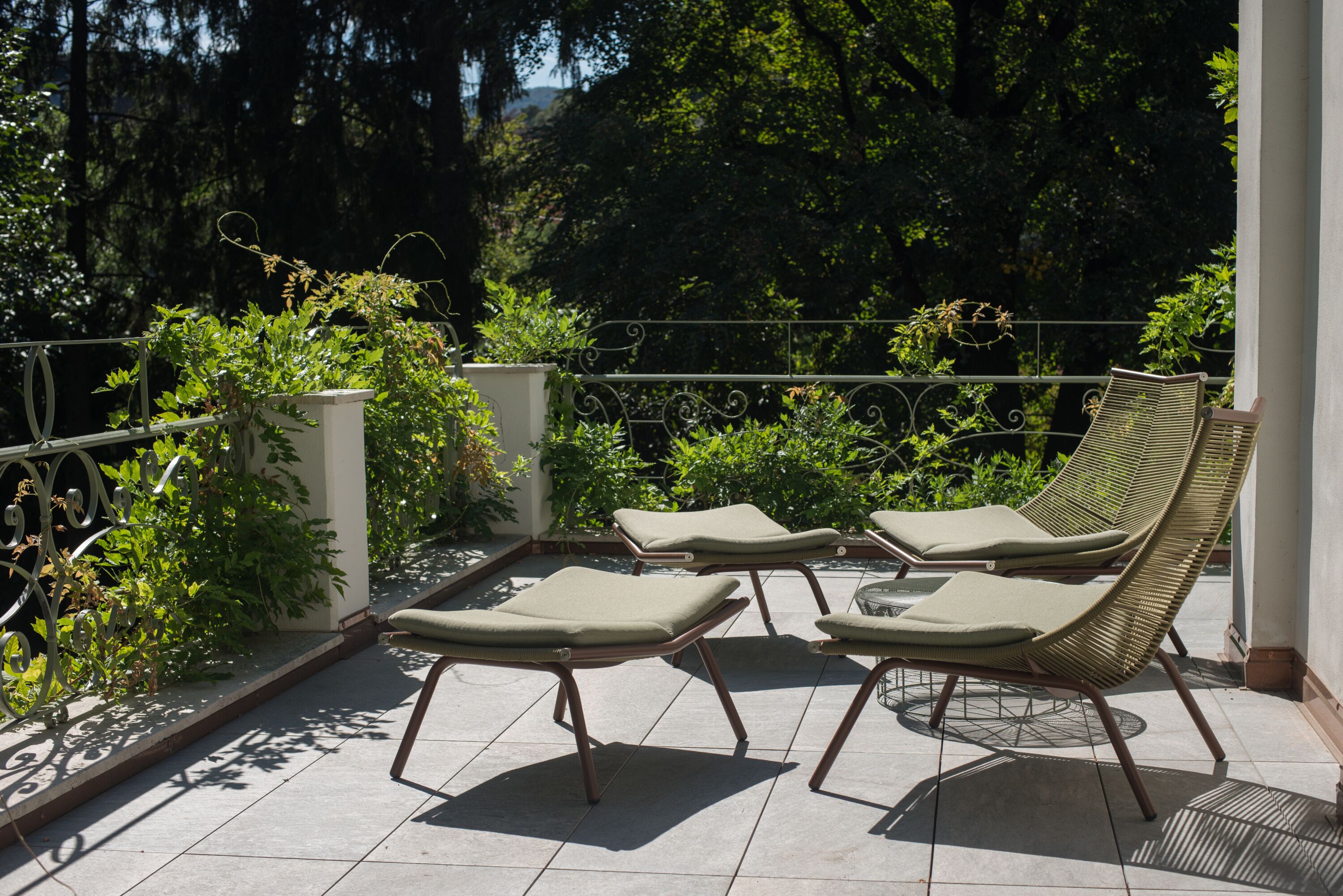 Une terrasse dotée de quatre fauteuils de jardin en osier et d’une petite table, entourée de verdure et d’une rambarde en métal.