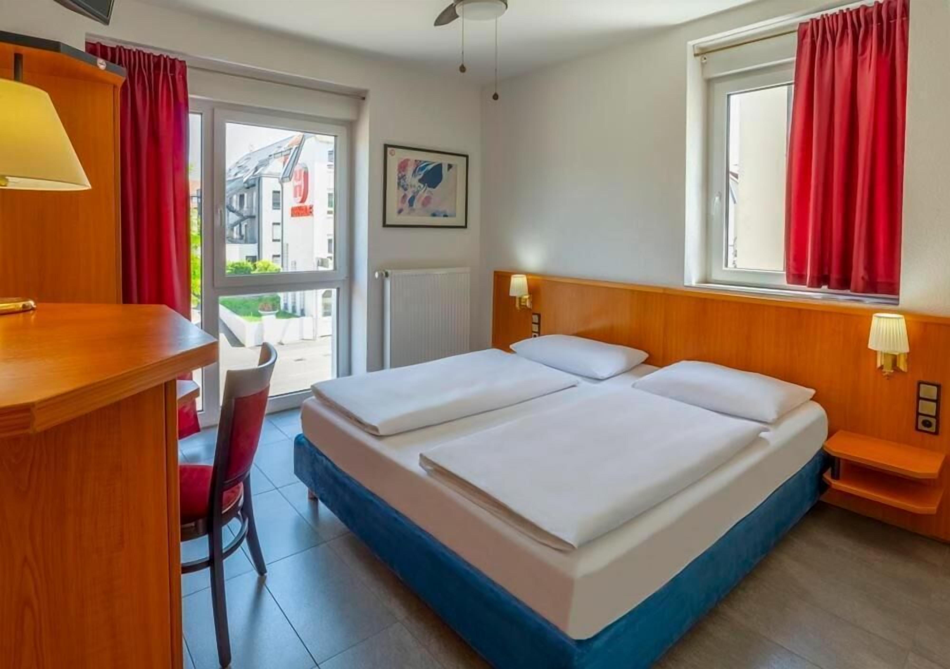 A hotel room with a double bed, a desk with a chair, a window with red curtains, and a view of buildings outside.