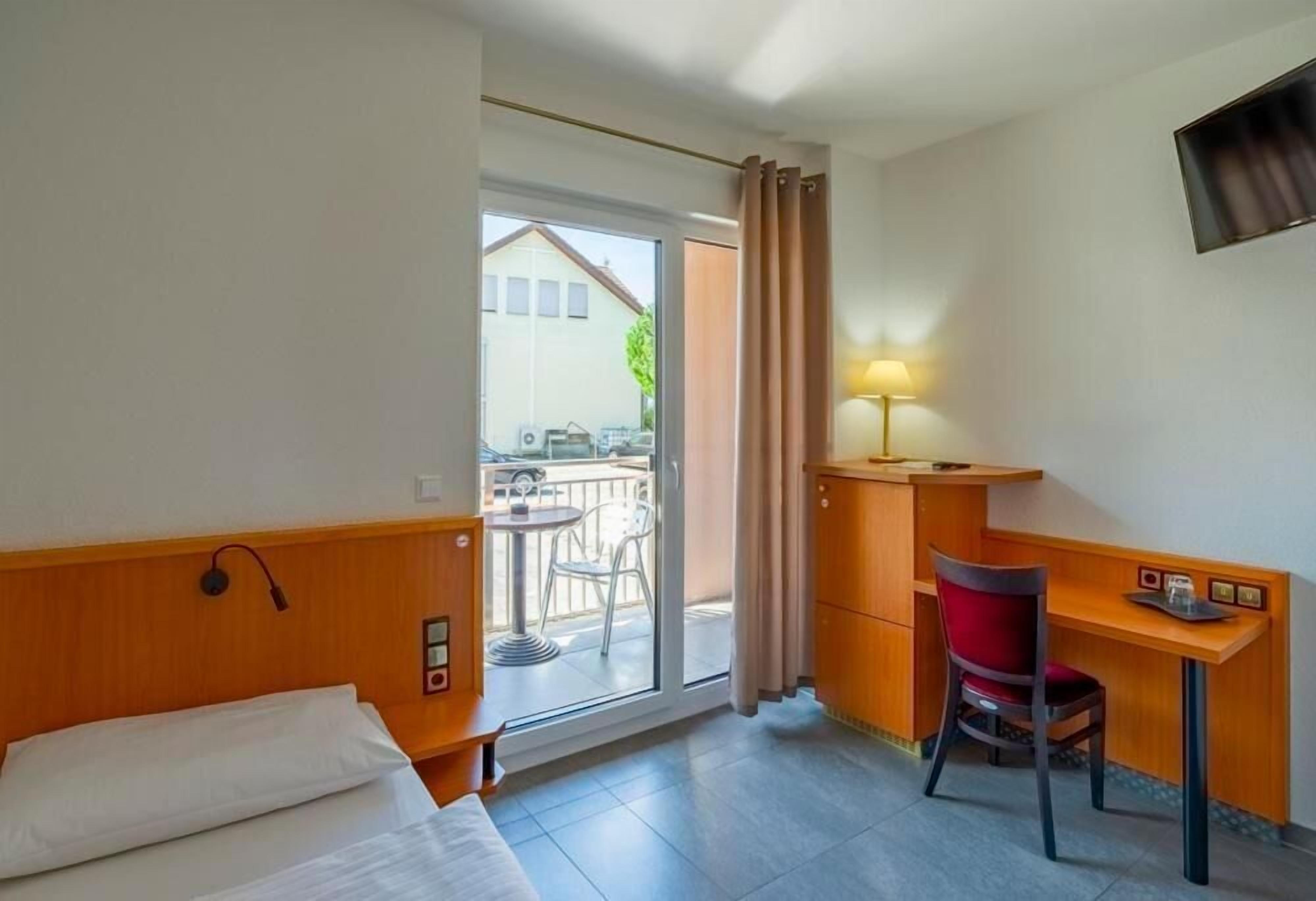 A hotel room with a bed, a desk with a chair, a lamp, and a view of the outside balcony.