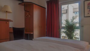 Hotellihuone, jossa on kaksi sänkyä, puinen vaatekaappi, työpöytä lampun kanssa ja ikkunasta avautuva näkymä ulos, ikkunassa on punaiset verhot.