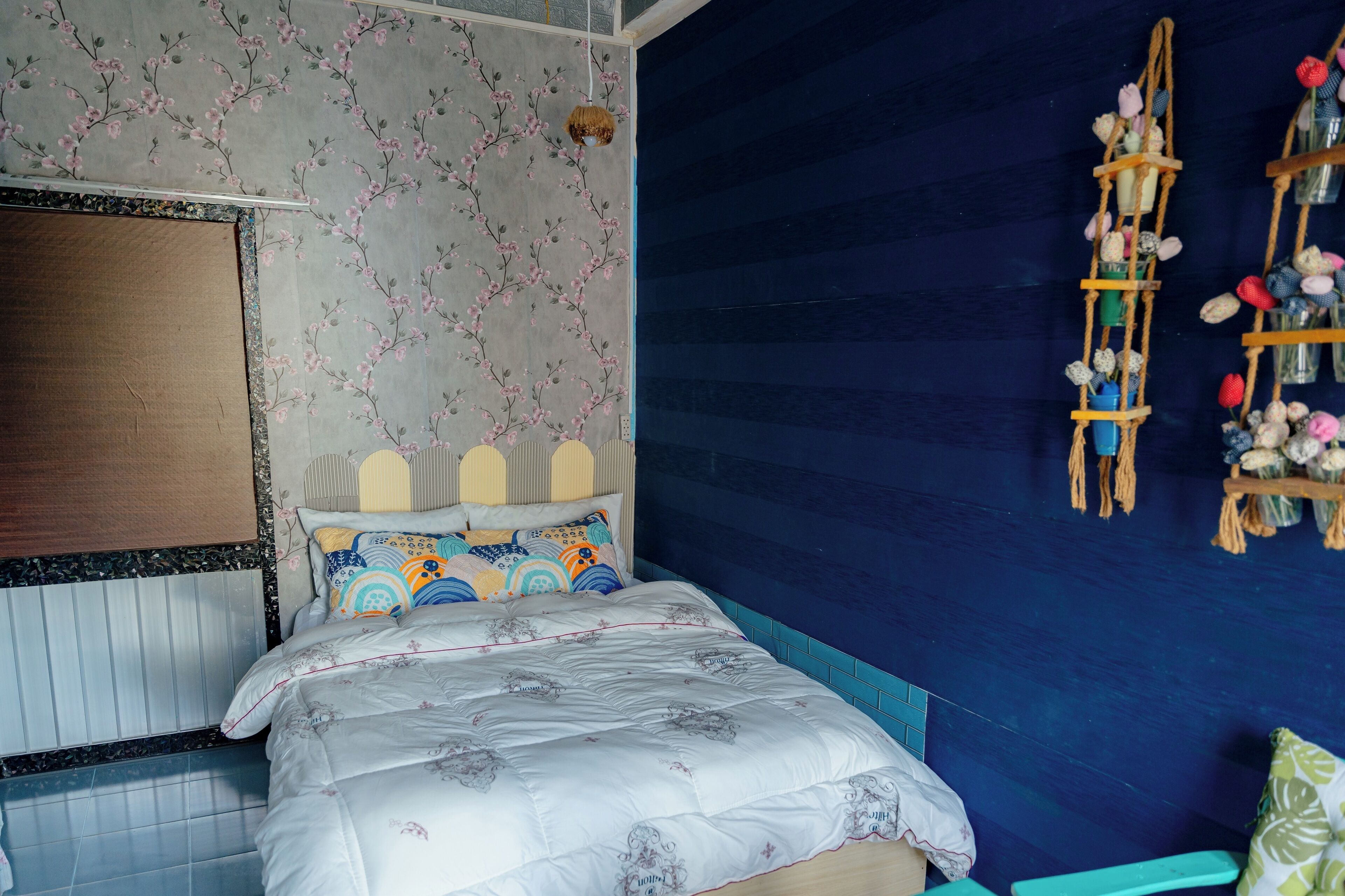 A bedroom with a bed, a wall-mounted shelf with flowers, and a patterned wallpaper.