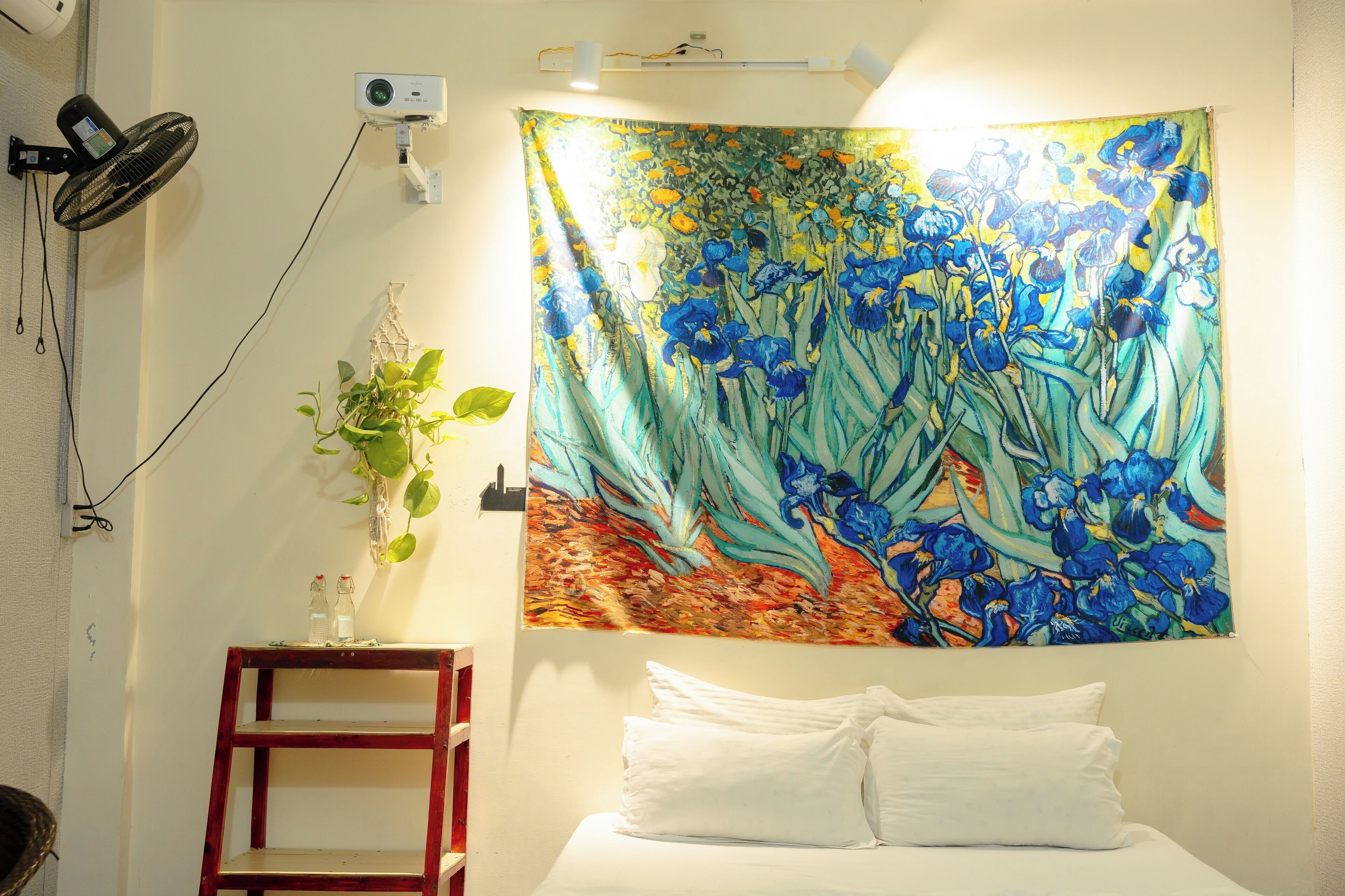 A bedroom with a bed, a tapestry with a floral design, a small table with a plant, and a wall-mounted projector.