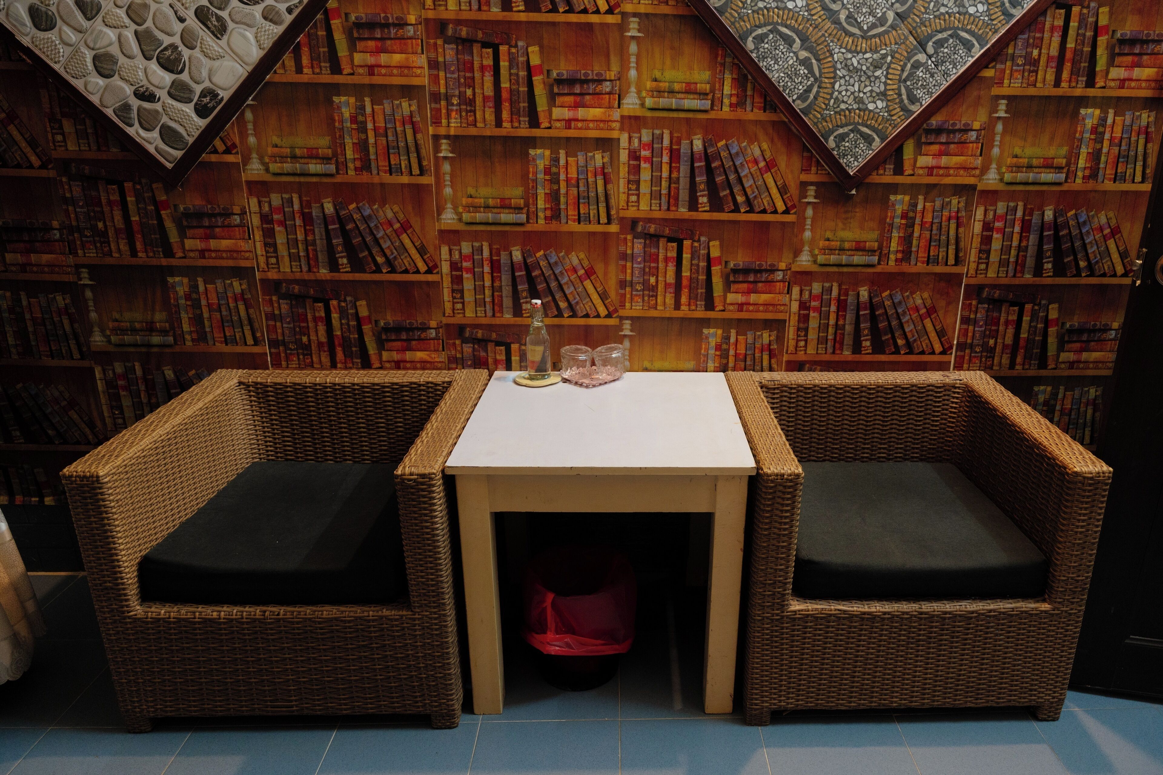 A room with two wicker chairs, a small table, and a bookshelf filled with books.