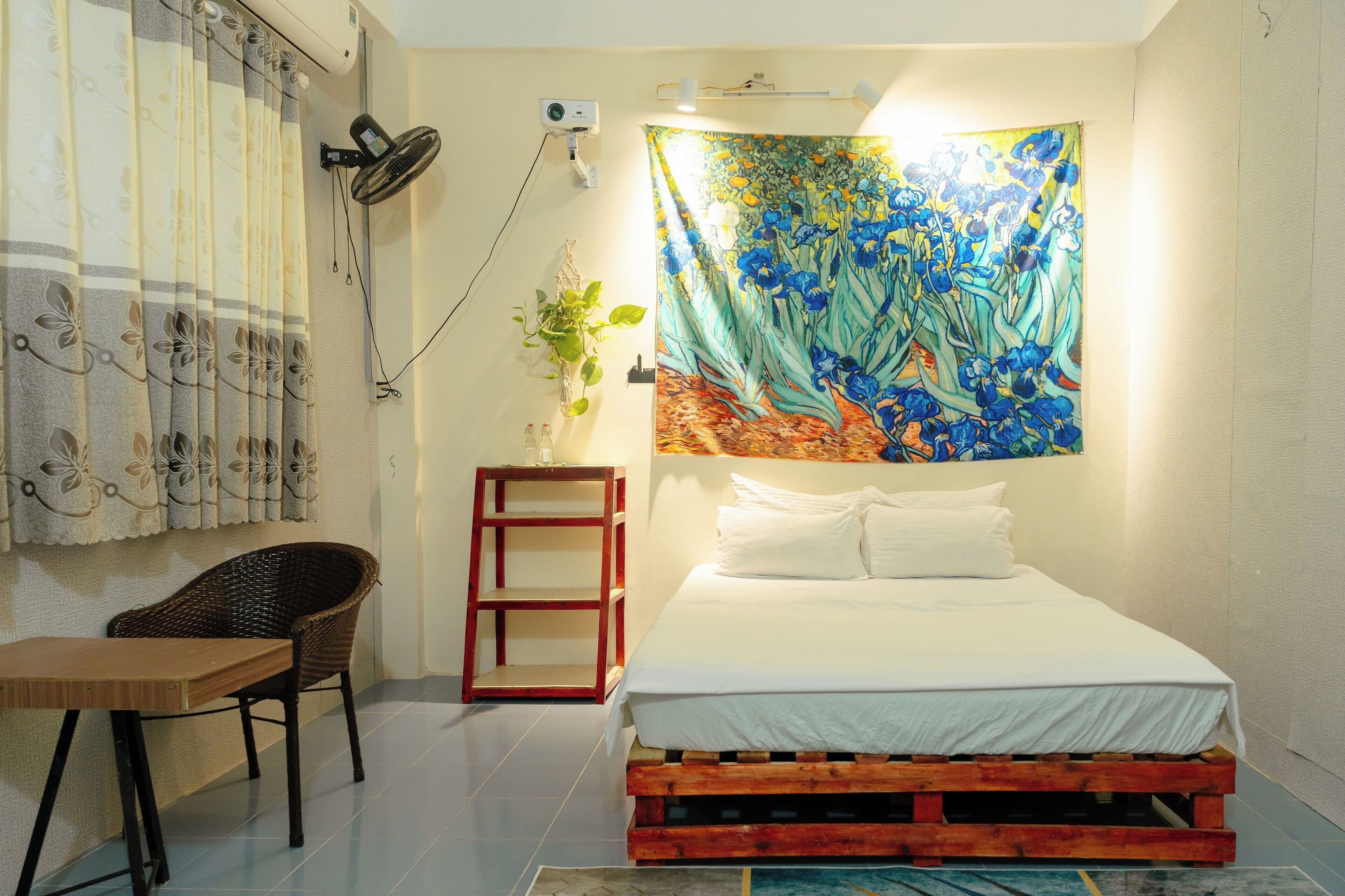 A bedroom with a bed, a chair, a small table, a tapestry, and a wall-mounted fan.