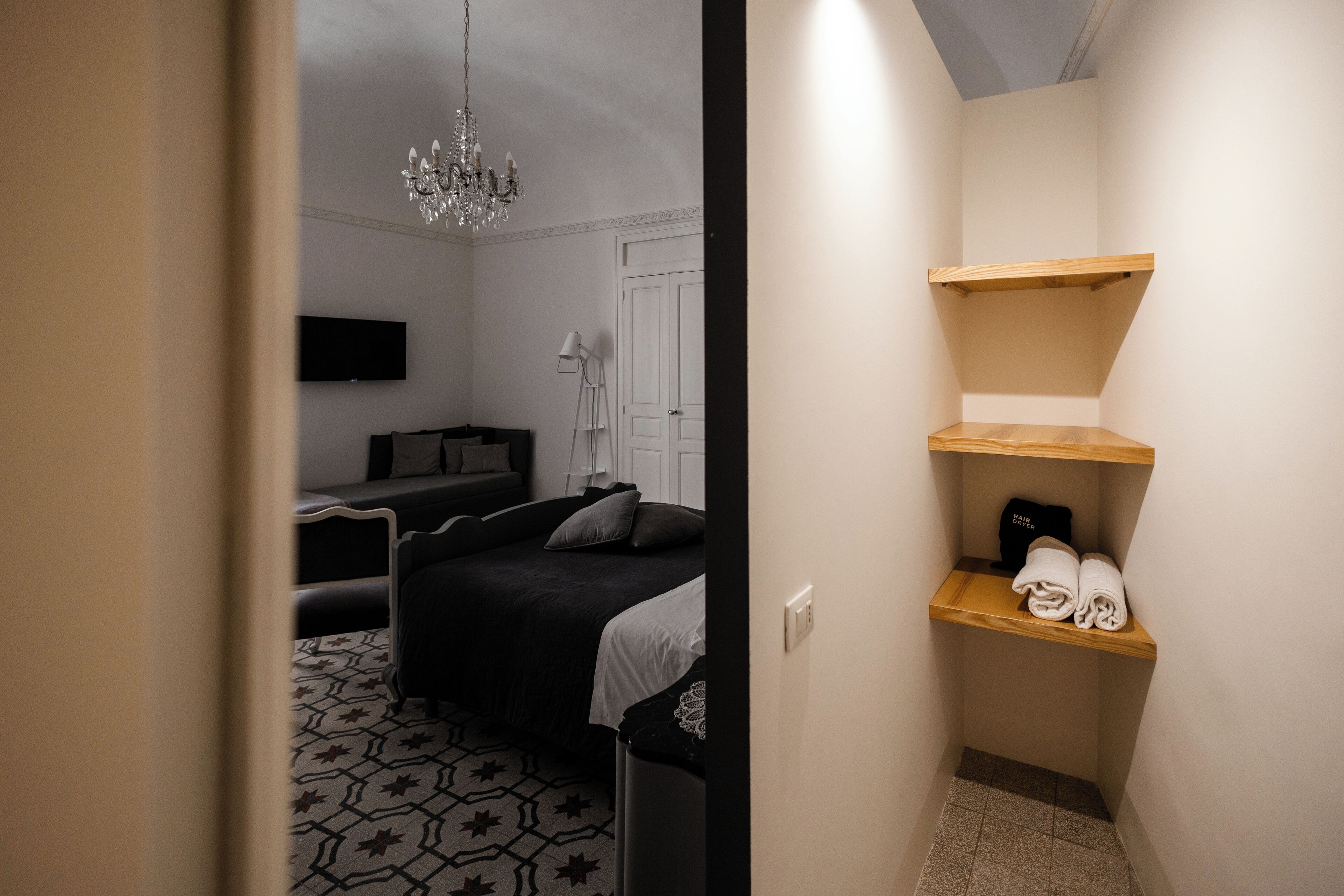 A compact hotel room with a bed, a TV mounted on the wall, a chandelier, and a small shelf with towels.