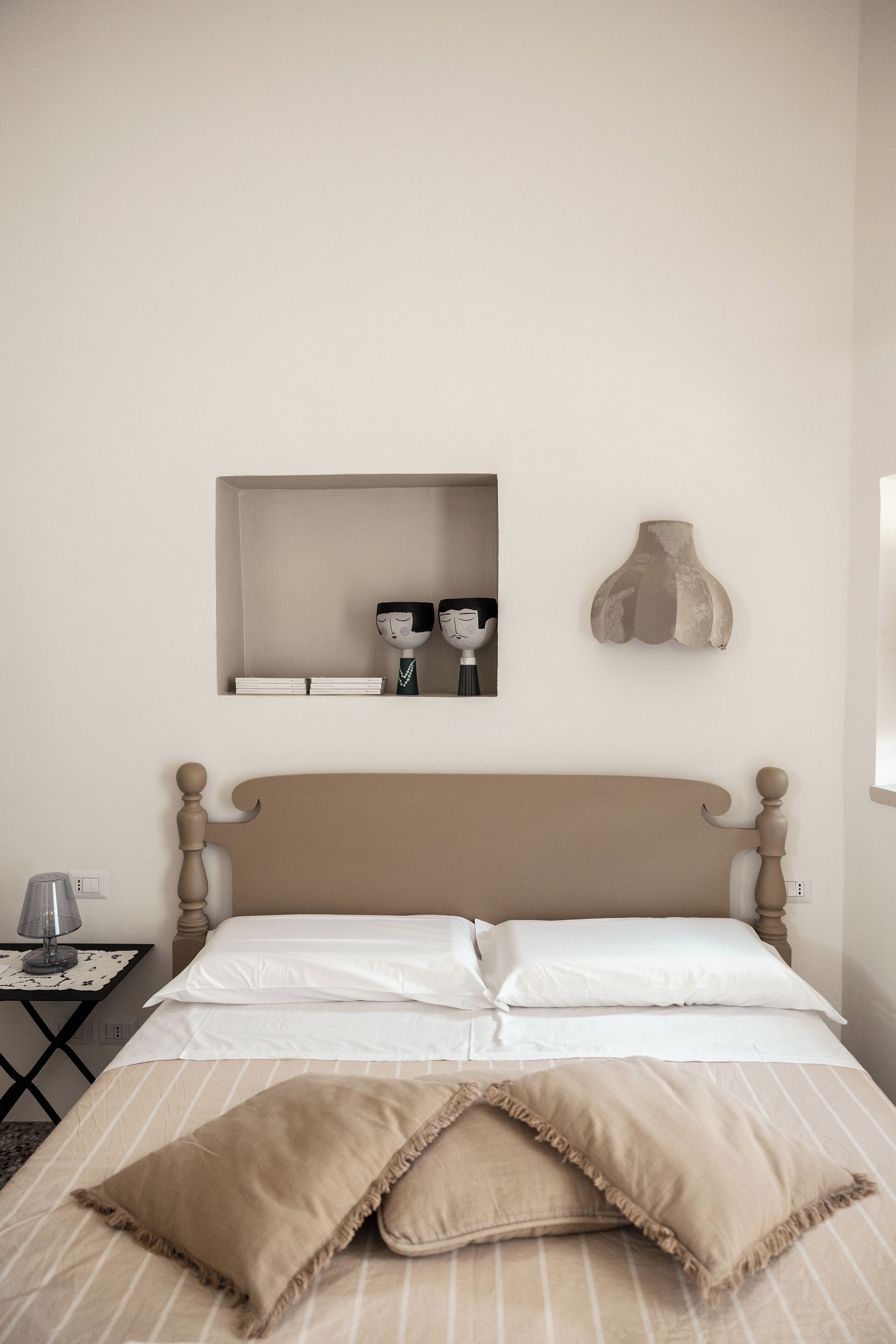 Una cama bien hecha con una colcha a rayas y dos almohadas, una mesita de noche con una lámpara, y un estante en la pared con objetos decorativos.