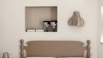 Una cama bien hecha con una colcha a rayas y dos almohadas, una mesita de noche con una lámpara, y un estante en la pared con objetos decorativos.