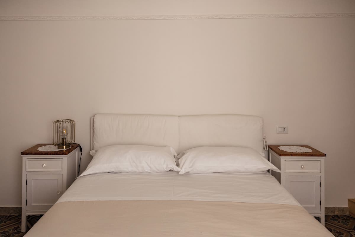 Una cama bien hecha con sábanas blancas, dos mesitas de noche con lámparas y una pared lisa.