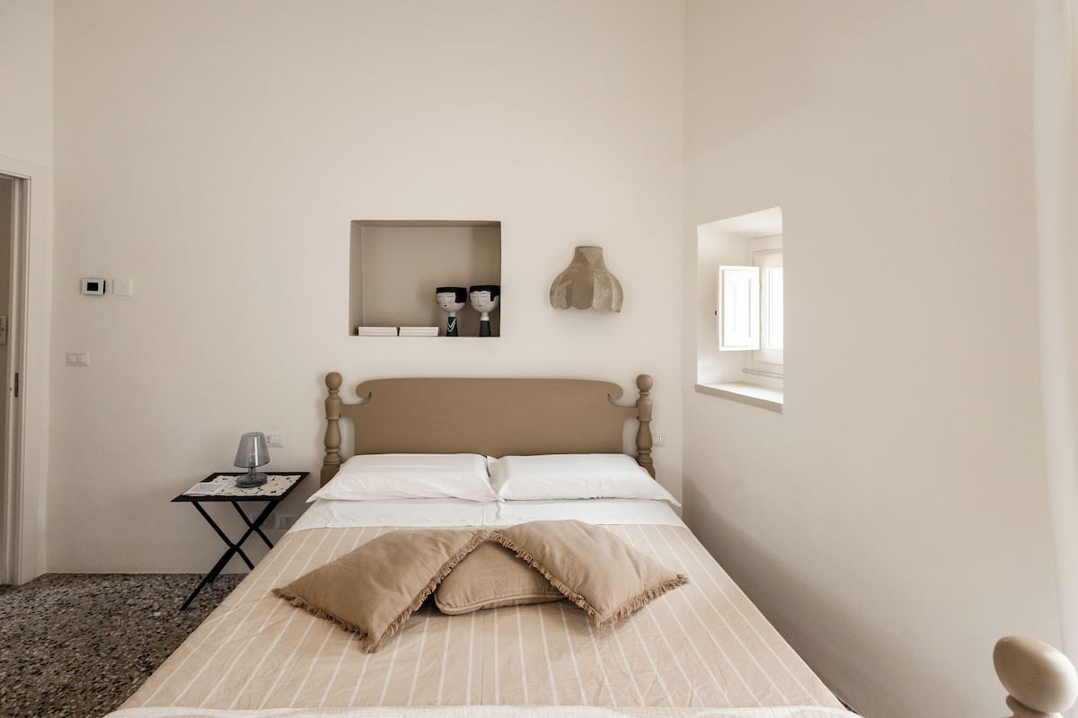 Un dormitorio con cama, mesita de noche con lámpara, estante pequeño con libros y espejo, y repisa en la pared con adornos.