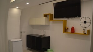 Una habitación moderna y bien iluminada, con un refrigerador blanco, un mueble negro, un televisor montado y un pequeño basurero.