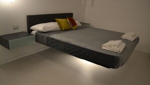 Una cama moderna con colchón gris, almohadas blancas y una almohada amarilla decorativa. Hay toallas blancas dobladas sobre la cama.
