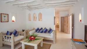 A room with a white sofa, a wooden coffee table, and a ceiling fan. There are arched windows and a door leading to another room. - Medina Palms (Watamu)