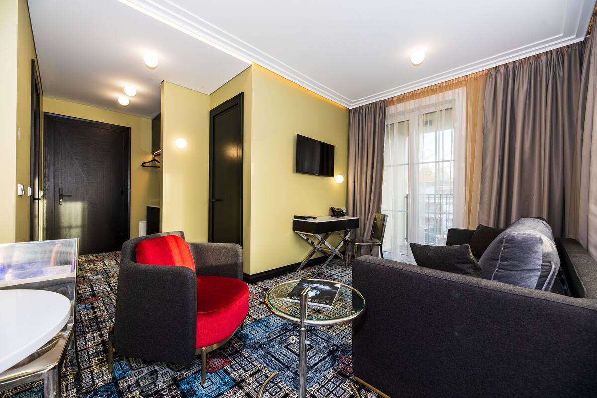 Een moderne hotelkamer met een bank, fauteuil en een klein tafeltje. Er is een televisie aan de muur bevestigd en een deur naar de badkamer.