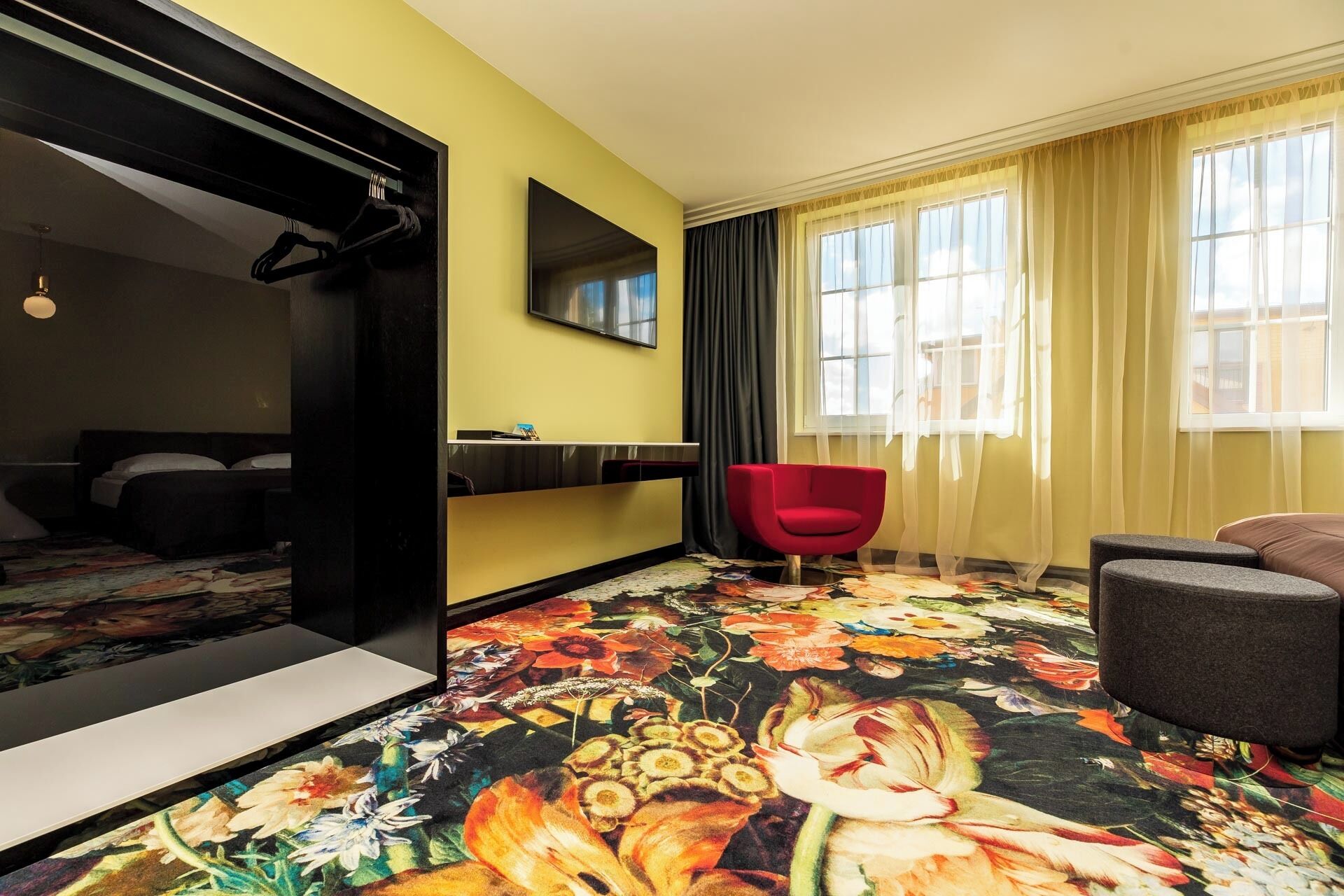 Habitación de hotel con una cama, una silla roja, un sofá gris, un espejo grande y un televisor.