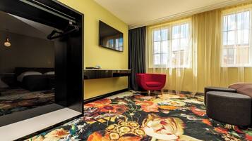 Habitación de hotel con una cama, una silla roja, un sofá gris, un espejo grande y un televisor.
