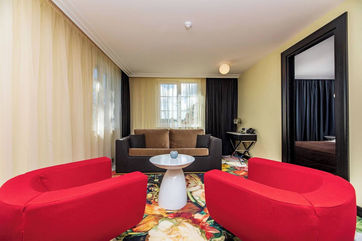 Een hotelkamer met een bank, twee rode stoelen, een klein tafeltje, een telefoon en een raam met gordijnen.