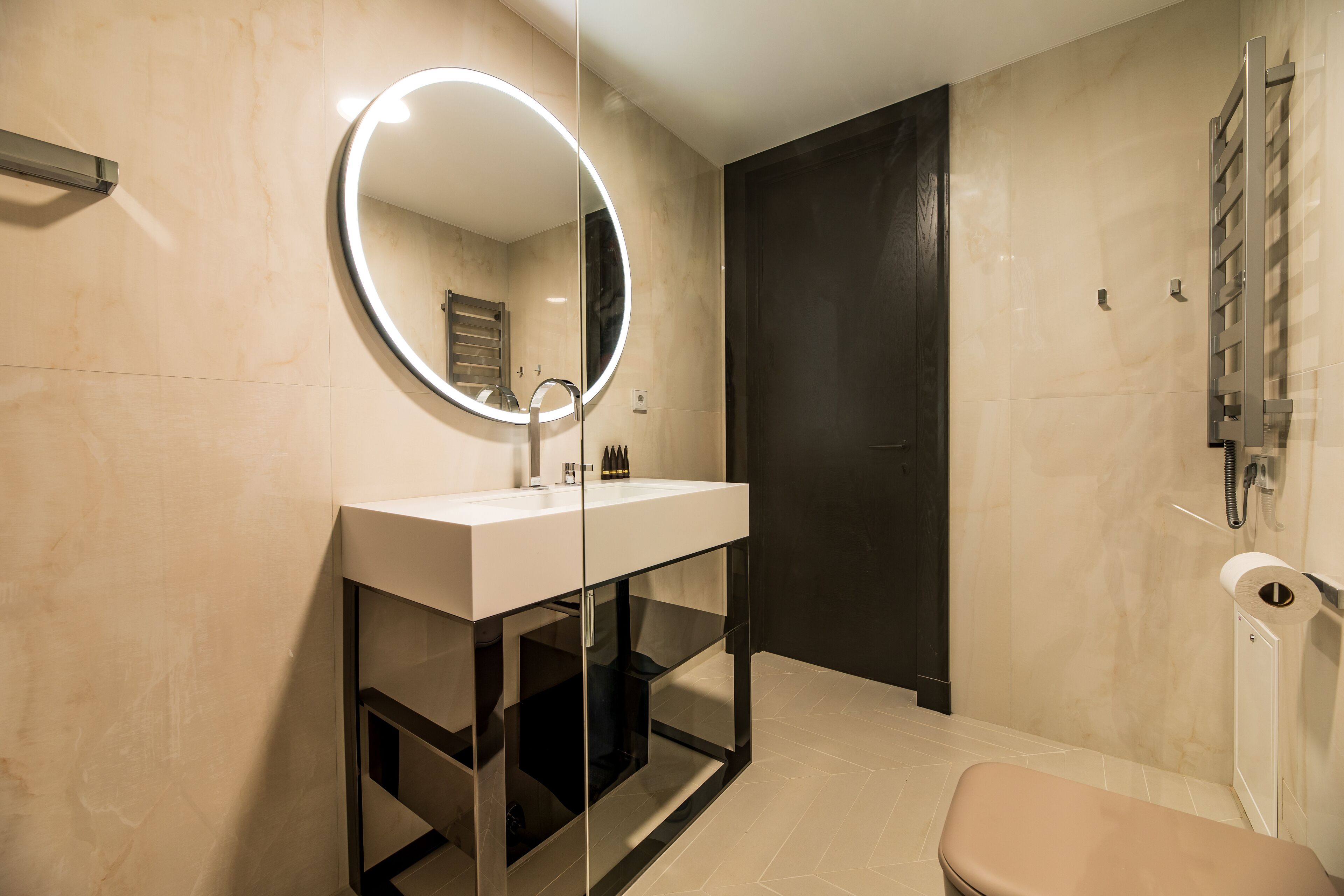 Een moderne badkamer met een glazen wastafelmeubel, een ronde spiegel met verlichting, een toilet en een donkere deur.