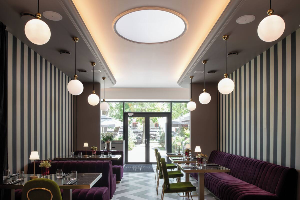 Een restaurant met gestreepte wanden, ronde plafondlampen en een centraal rond dakvenster. Tafels en stoelen staan klaar voor het diner.