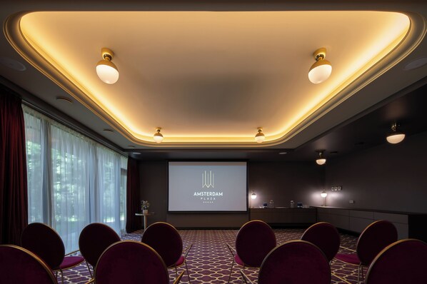 Un salón de conferencias con una pantalla grande que muestra “AMSTERDAM PLAZA”, filas de sillas rojas, piso alfombrado e iluminación empotrada en el techo.