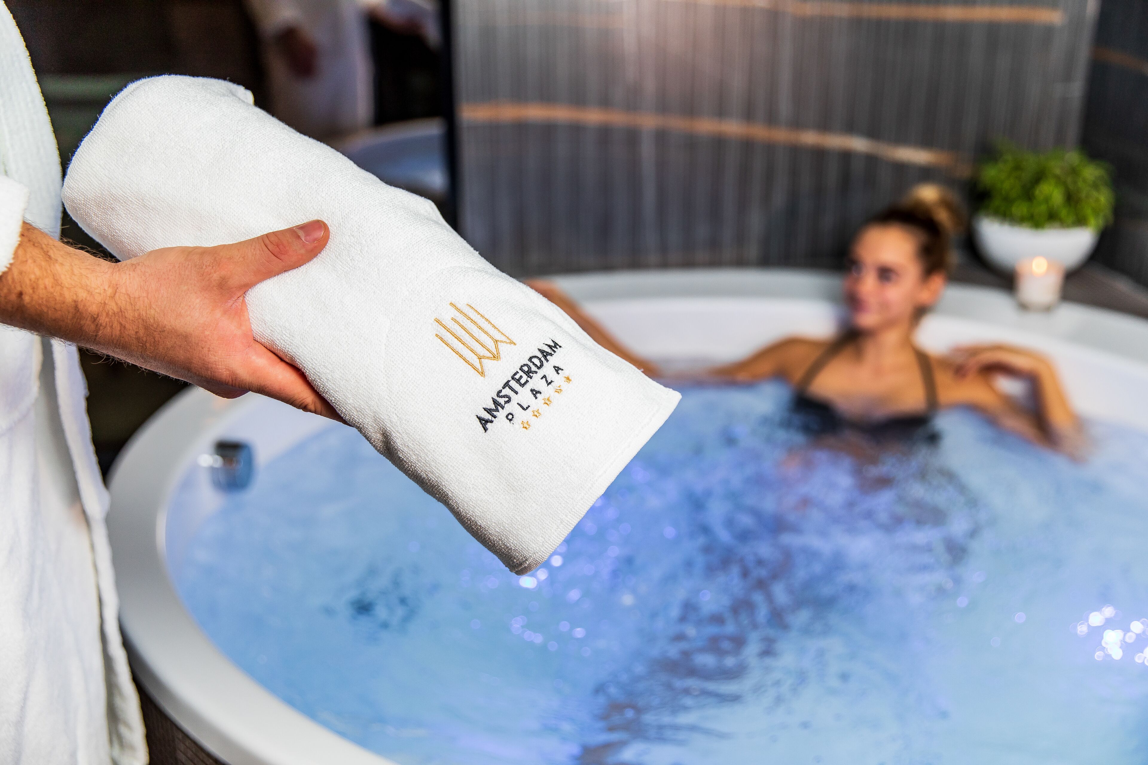 Una persona sosteniendo una toalla con el texto "AMSTERDAM SPA & PLAZA", una bañera de hidromasaje con agua y una planta al fondo.