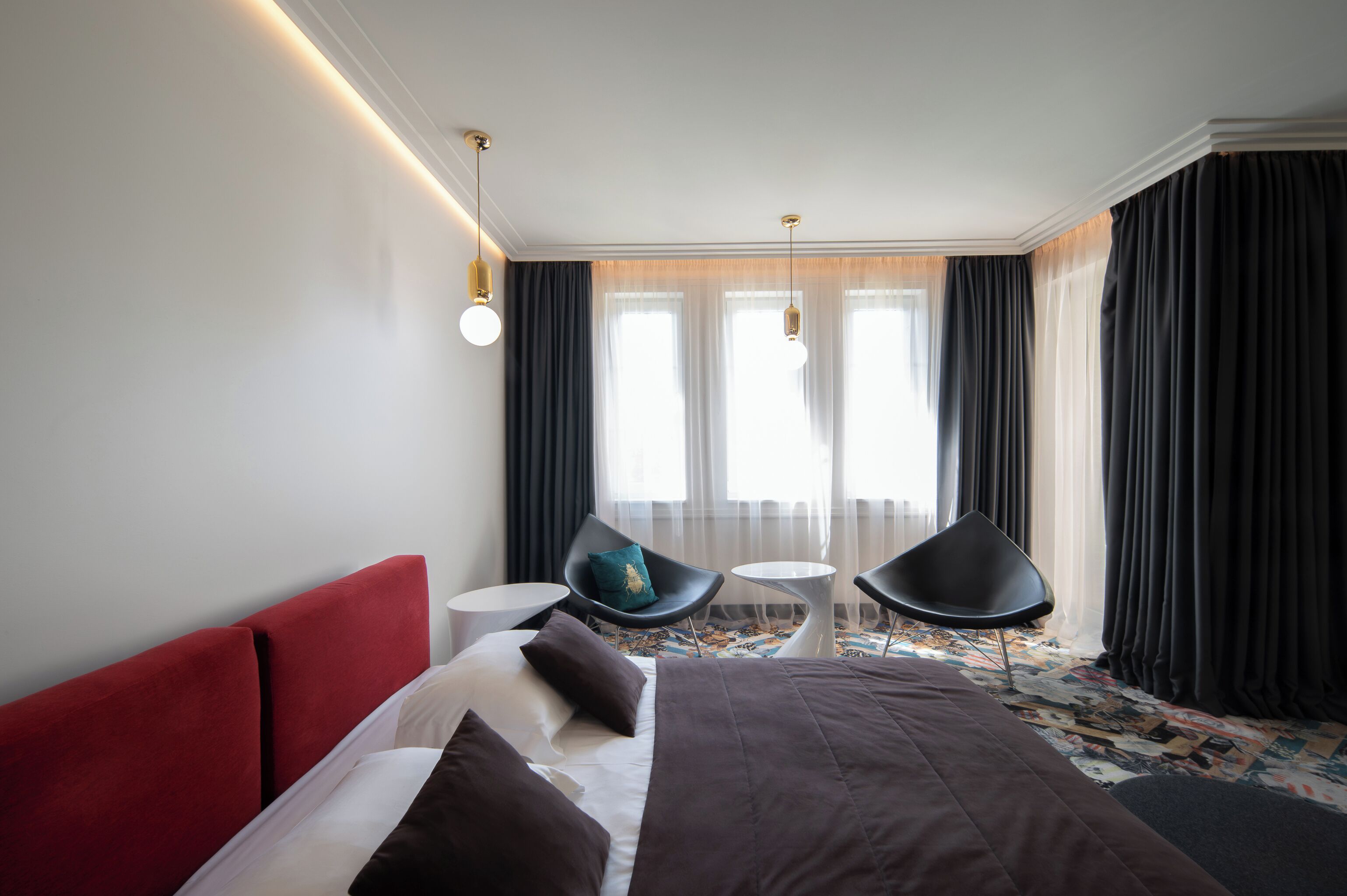 Une chambre d’hôtel moderne, dotée d’un grand lit, de deux fauteuils, d’une petite table et offrant une vue sur la ville grâce à de grandes fenêtres.