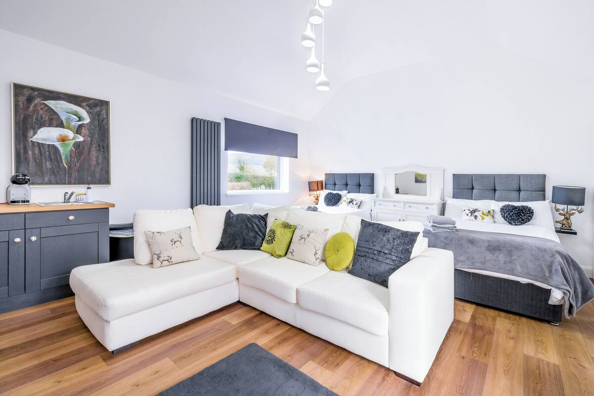 A modern bedroom with a sectional sofa, a bed with a grey headboard, a bedside table, and a window with curtains.
