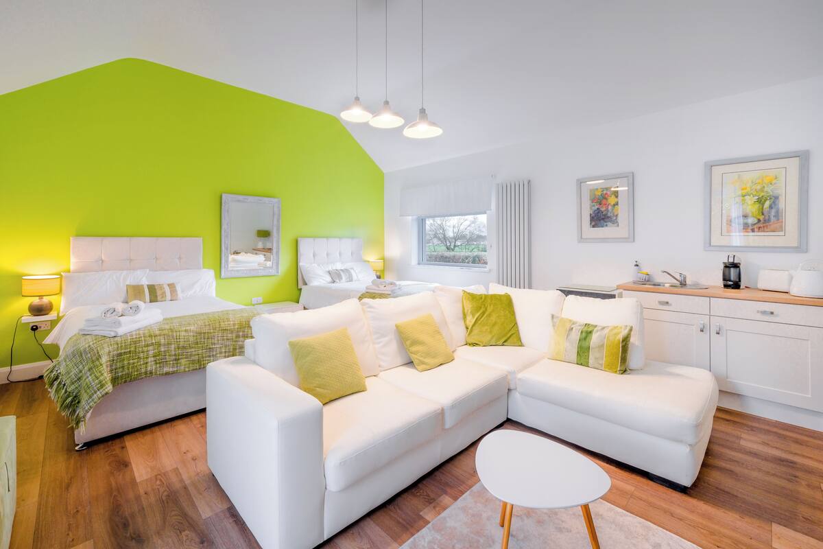 A modern living room with a white sofa, a green accent wall, and a kitchen area with white cabinets.