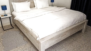 Una cama bien hecha con sábanas y almohadas blancas, una mesita de noche con una lámpara y un cuadro en la pared.