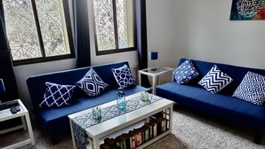 Un salón con sofá azul, cojines estampados, una mesa de centro con libros y vistas a través de una ventana con barrotes.