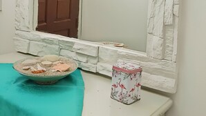 Una habitación con pared blanca con textura, un espejo grande, una puerta de madera, un mantel color esmeralda, un cuenco con adornos y un recipiente estampado con motivos florales.