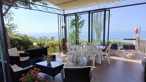 Eine überdachte Terrasse mit Korbmöbeln, einem Tisch mit Blumen in einer Vase und Blick aufs Meer.