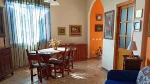 Ein Essbereich mit Tisch, Stühlen und einer Blumenvase. Im Vordergrund befindet sich ein blaues Sofa und eine Lampe.