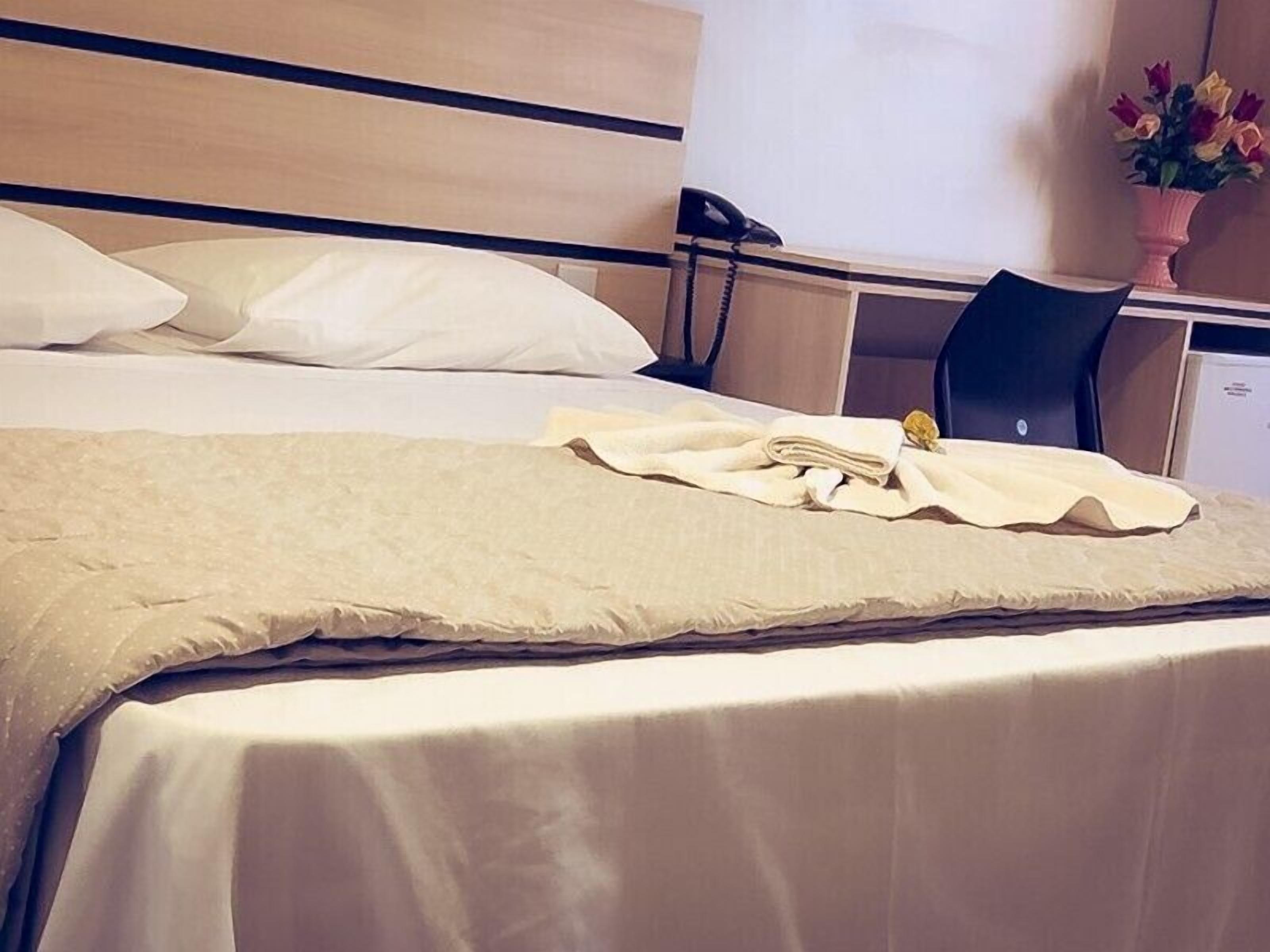 Una cama bien hecha con una manta y almohadas color beige, un teléfono en la mesita de noche, un pequeño escritorio con una silla y un florero con flores.