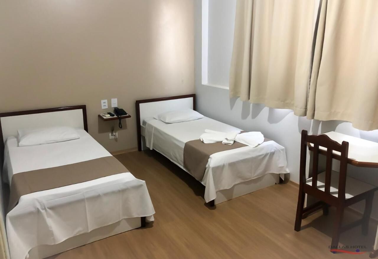 Quarto de hotel com duas camas de solteiro, uma cadeira de madeira, uma mesa pequena e uma janela com cortinas.