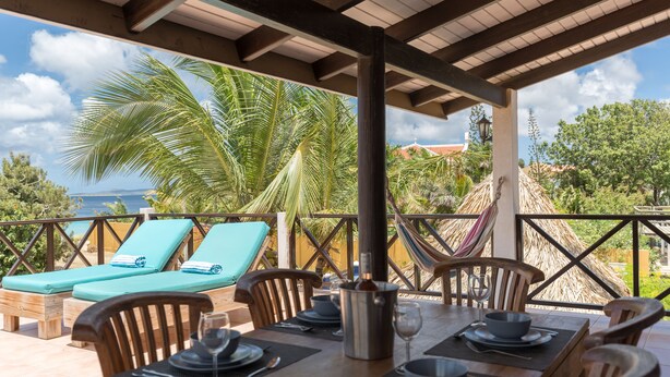 Un área de comedor al aire libre techada con una mesa lista para dos personas, sillones de descanso y vista al océano.