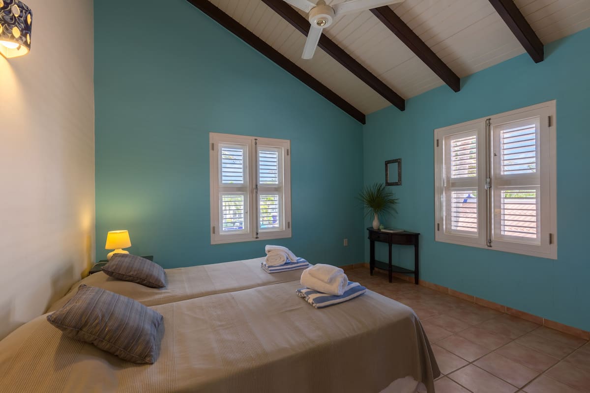 Una habitación con dos camas, ventilador de techo y dos ventanas con contraventanas.