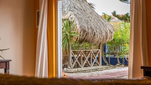 Vista a través de un umbral hacia un espacio tropical al aire libre con una estructura de techo de paja, una palmera y una cerca de madera.