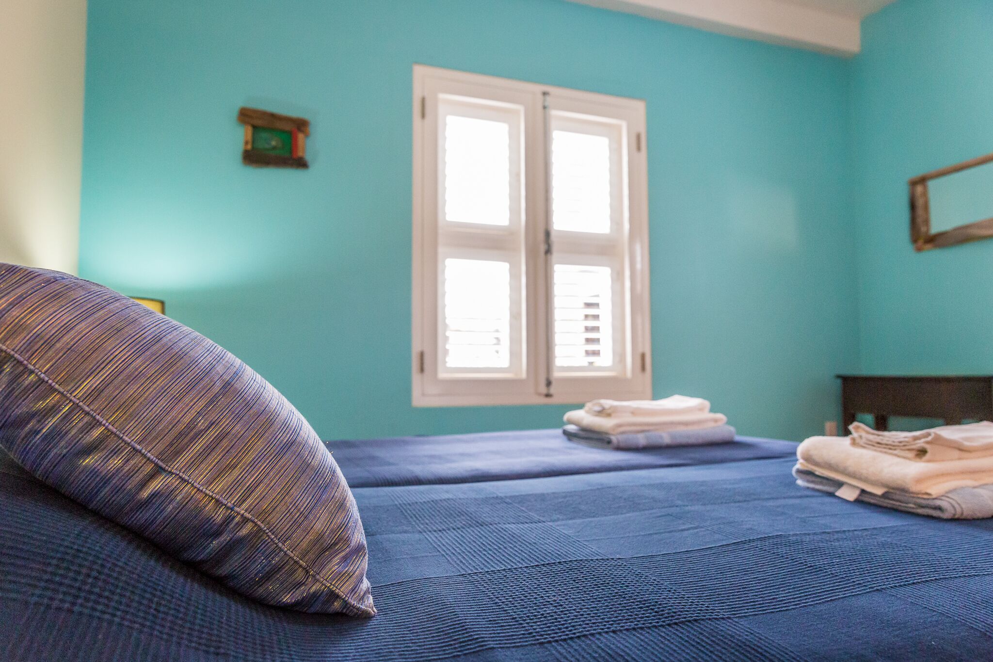 Ein Zimmer mit türkisfarbenen Wänden, einem Bett mit blauer Bettdecke, einem Fenster mit weißen Lamellen und gefalteten Handtüchern auf dem Boden.