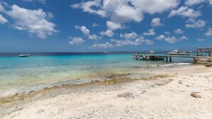 Una playa con agua turquesa cristalina, un muelle de madera y barcos a lo lejos.