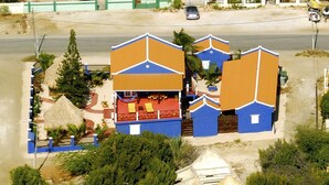 Vista aérea de una propiedad costera con varias edificaciones, incluyendo una estructura grande con fachada azul y naranja, un edificio más pequeño con techo rojo, y una casa con techo de paja. Se observa una zona de playa arenosa con vista despejada al mar turquesa.