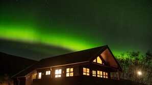 Uma cabana de madeira com várias janelas, sob um céu estrelado com aurora boreal visível.