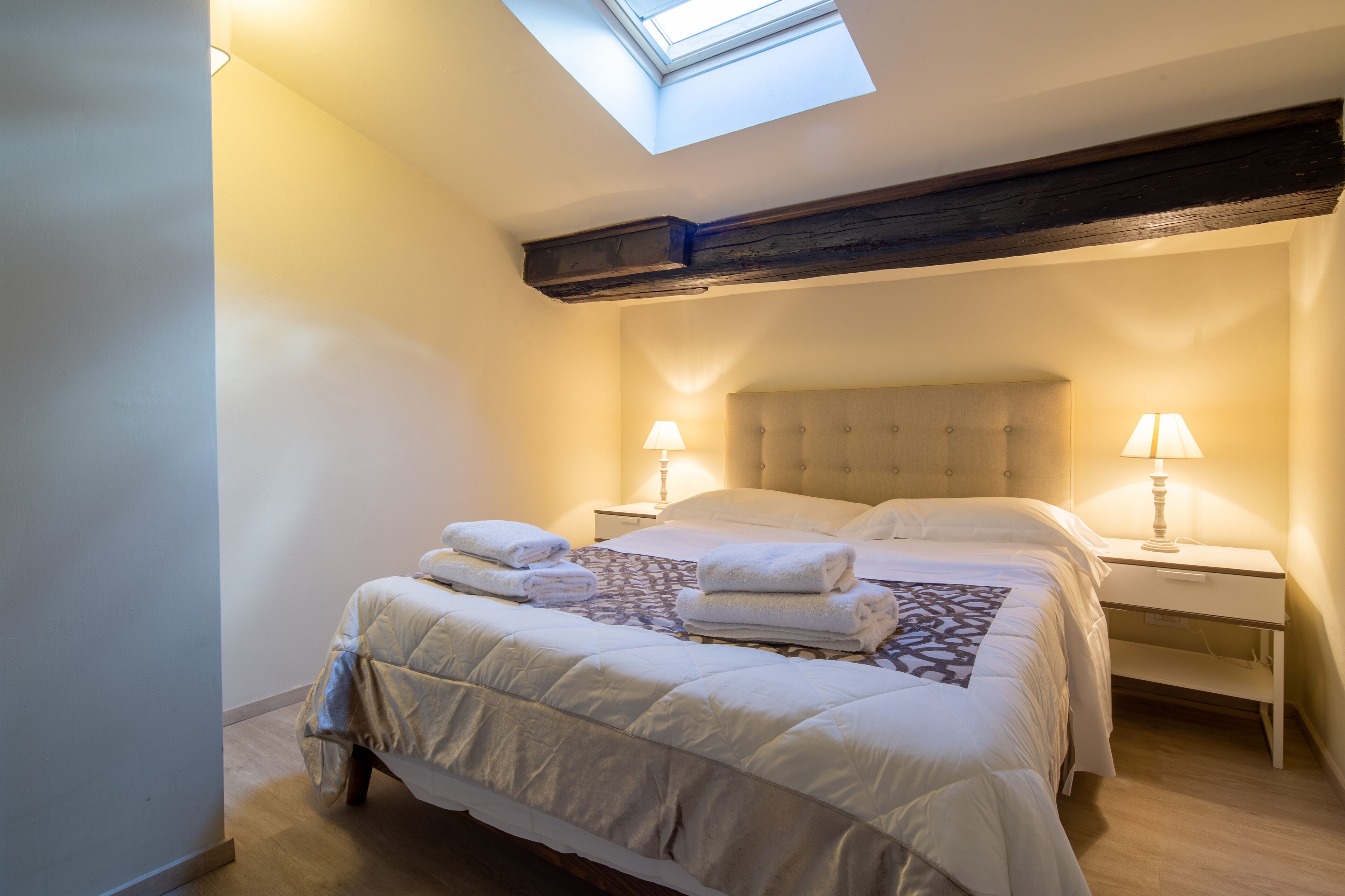 A bedroom with a skylight, a large bed with multiple pillows and a blanket, bedside tables with lamps, and a wooden beam above the bed.
