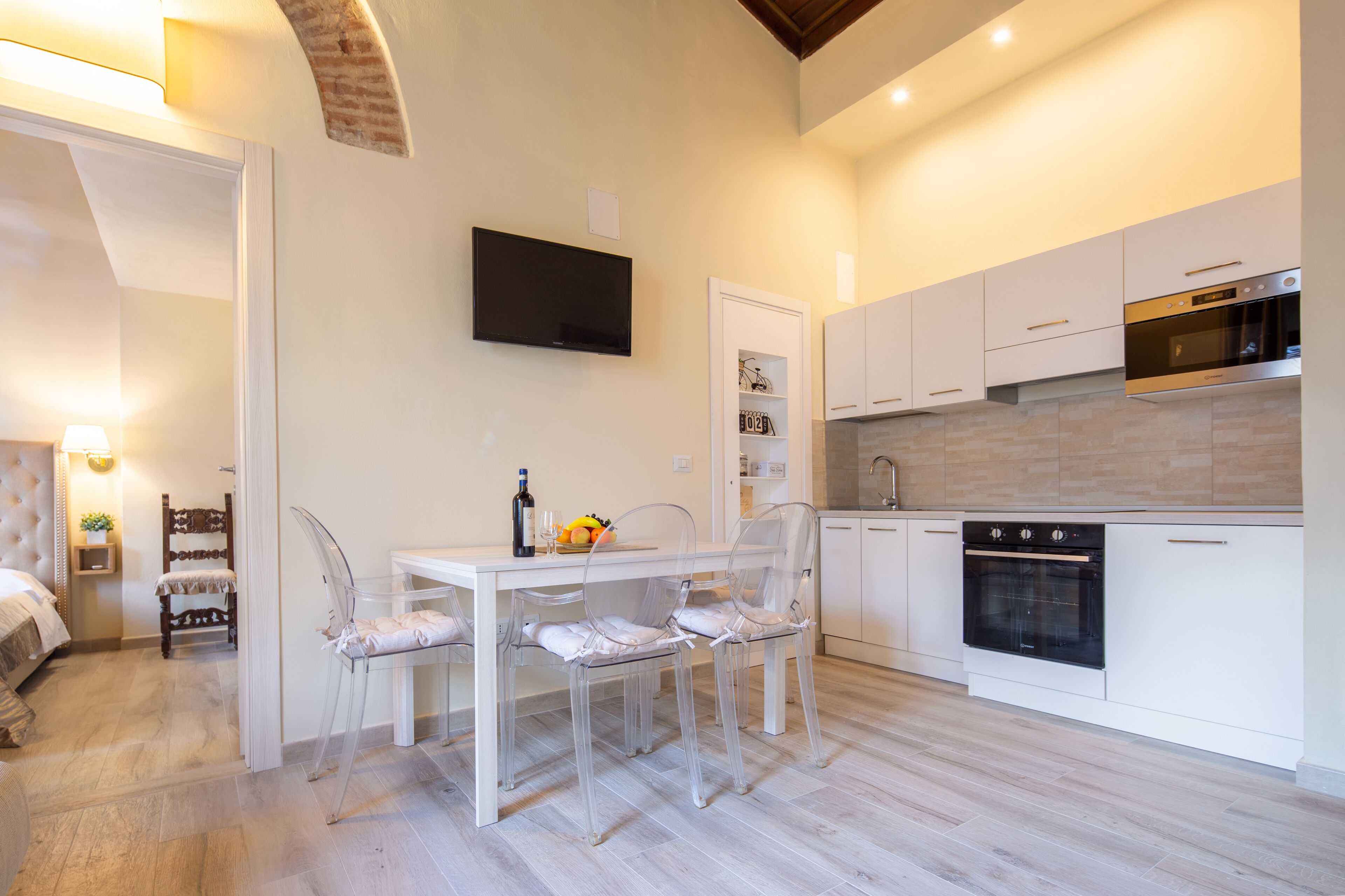 Una cocina moderna con armarios blancos, horno empotrado y comedor con mesa blanca y sillas transparentes.