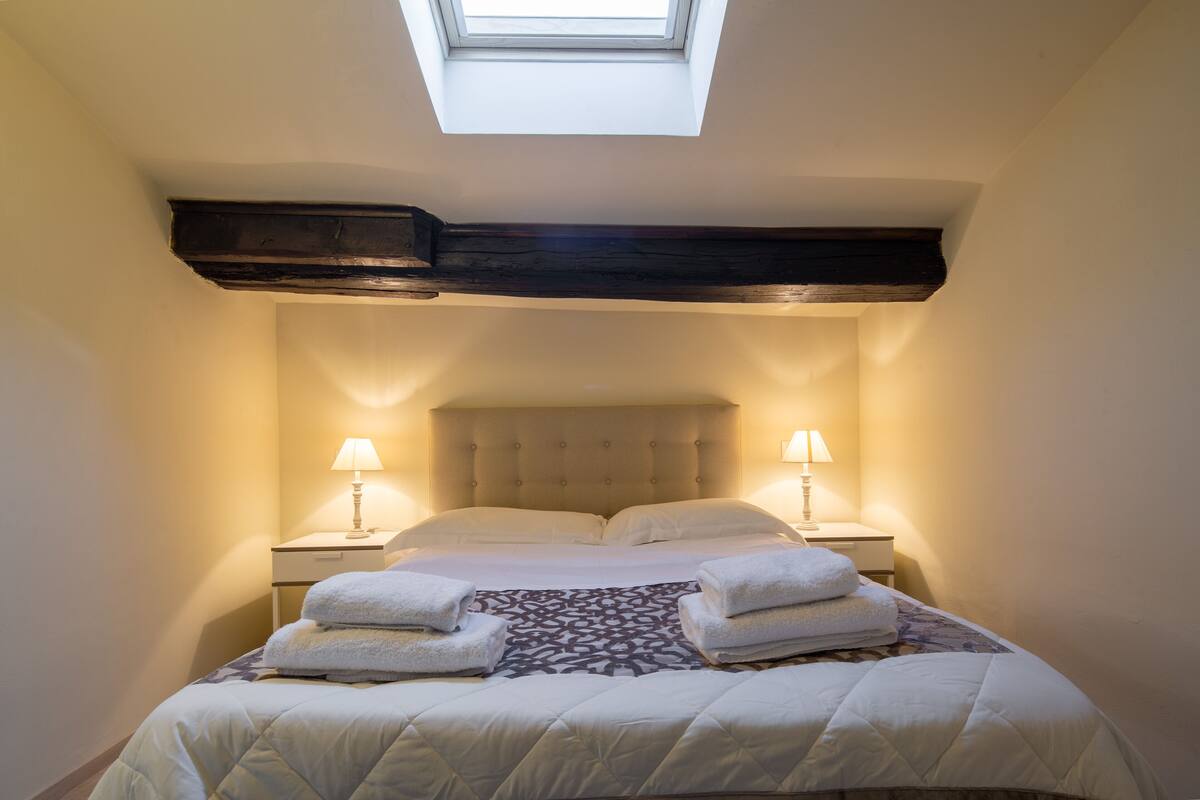 A bedroom with a skylight, two bedside lamps, a bed with a patterned bedspread, and two towels on the bed.