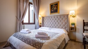 A hotel room with a large bed, a headboard, two bedside lamps, a window with curtains, and a painting on the wall. - Duomo Apartments by Mmega (Florence)