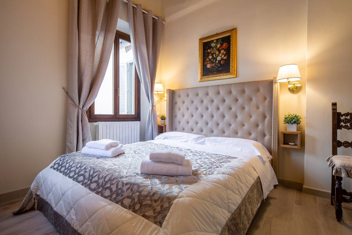 A hotel room with a large bed, a headboard, two bedside lamps, a window with curtains, and a painting on the wall.