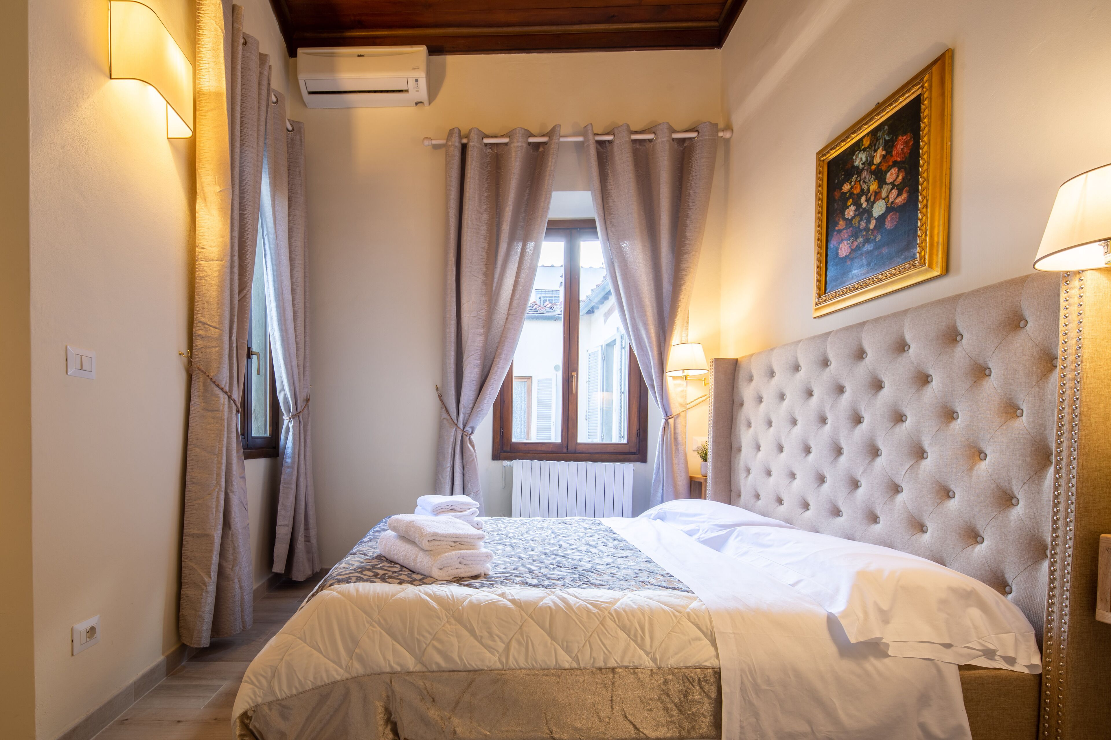 Habitación de hotel con una cama grande, cabecera capitonada, ventana con cortinas y un cuadro en la pared.