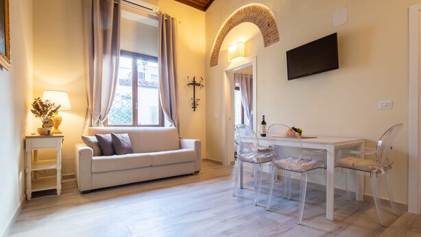 A living room with a sofa, a dining table with chairs, a TV mounted on the wall, and a window with curtains. - Duomo Apartments by Mmega (Florence)