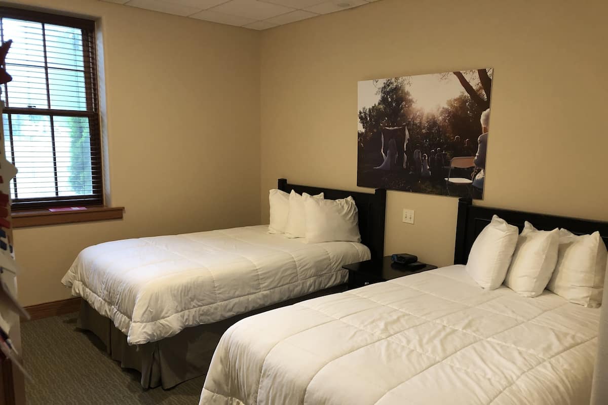 Ein Hotelzimmer mit zwei Einzelbetten, einem Fenster mit Jalousien, einem Bild an der Wand und einem kleinen Tisch zwischen den Betten.