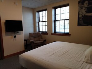 A hotel room with a bed, a chair, a television mounted on the wall, and windows with blinds.